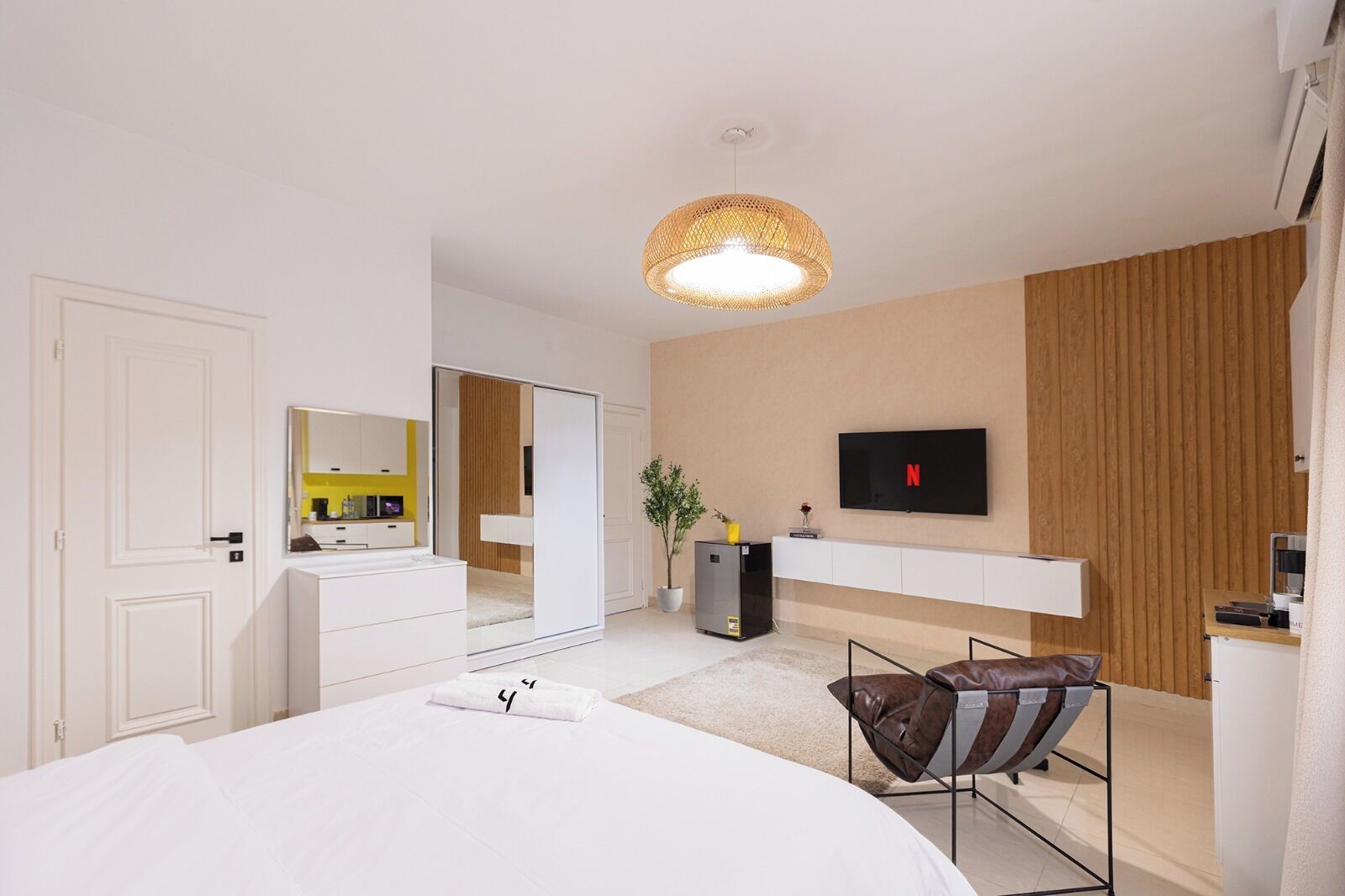 Quarto moderno com cama grande, televisão na parede, guarda-roupa de correr e um pequeno espaço de estar com uma poltrona.