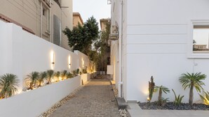 Un estrecho sendero empedrado flanqueado por paredes blancas, bordeado de pequeñas plantas en macetas e iluminado por luces fijadas en la pared.