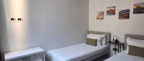 A hotel room with two single beds, a small table, a shelf with towels, and a wall with framed pictures.