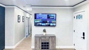 Un pasillo con una televisión instalada que muestra la interfaz de un servicio de streaming, una consola de madera con una planta y una puerta con diseño geométrico.