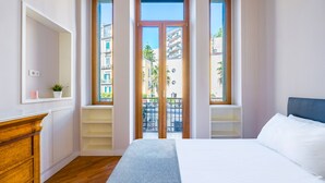 Une chambre avec un grand lit, une commode en bois et une vue sur les immeubles à travers une porte vitrée.