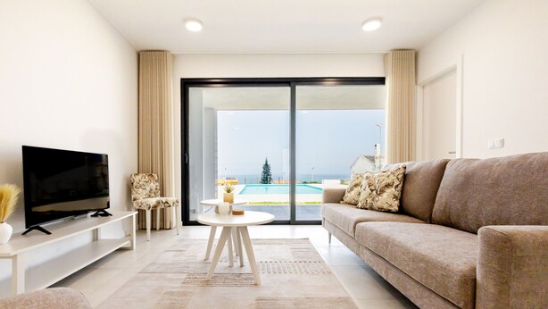A modern living room with a flat-screen TV, a beige sofa, a coffee table, and a view of a swimming pool through large glass doors. - Vale Furado Sea & Nature - BY SCH (Nazaré)