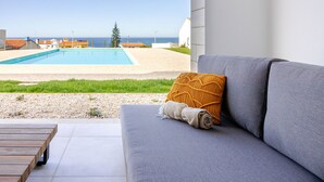 A modern outdoor seating area with a grey sofa, a wooden bench, and a view of a swimming pool and beach. - Vale Furado Sea & Nature - BY SCH (Nazaré)
