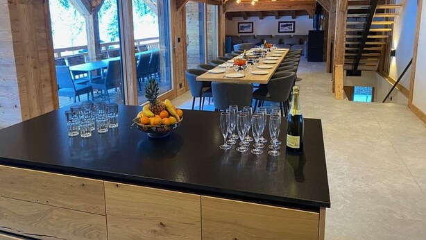 Un comedor moderno con una larga mesa dispuesta para una comida, un frutero en la encimera y una botella de champán.