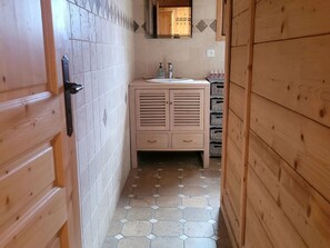 Een smalle gang met een houten deur, betegelde vloer, een houten kastje met spiegel en een wastafel.