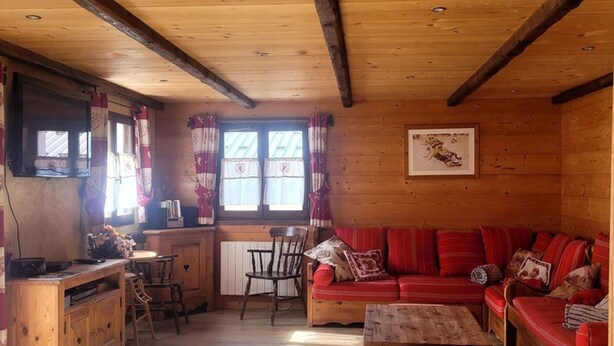 Indretning i en hyggelig hytte med træloft, sofaarrangement, spisebord og et køkken.