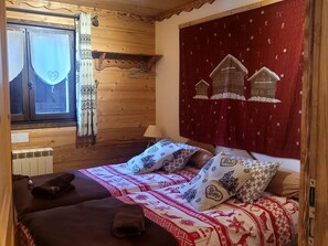 Een gezellige kamer met twee bedden, houten wanden, een rode wandtapijt met huisjes en een raam met gordijnen.
