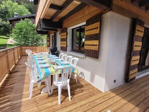 An outdoor dining area with a wooden deck, white plastic chairs, and a table set for a meal. - Renovated family chalet, near center, sleeps 12, parking, WiFi (Les Gets)