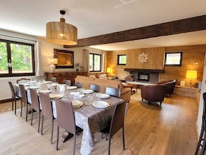 A dining area with a long table set for six, a fireplace, and a large window with a view of greenery. - Renovated family chalet, near center, sleeps 12, parking, WiFi (Les Gets)
