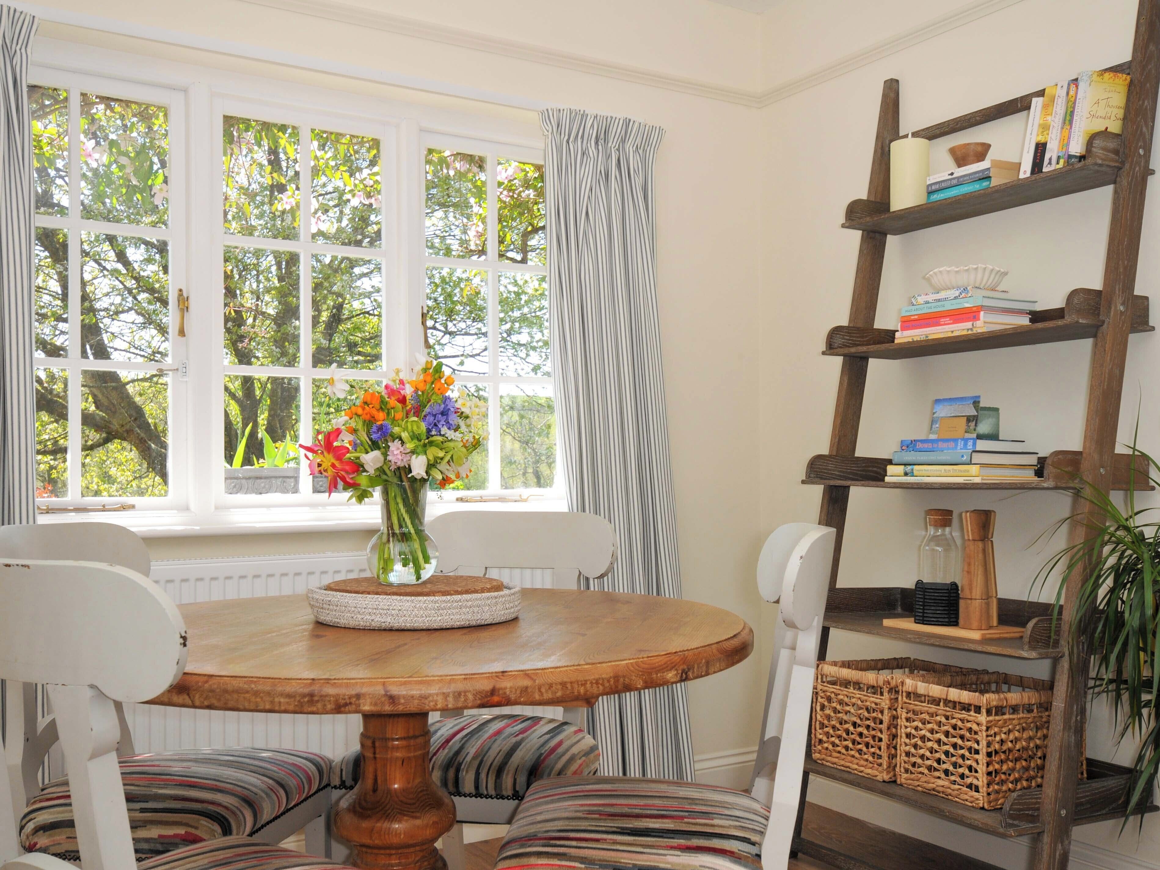 Un coin repas avec une table ronde en bois, des chaises à motifs rayés, un vase de fleurs et une bibliothèque contenant des livres et des objets décoratifs.