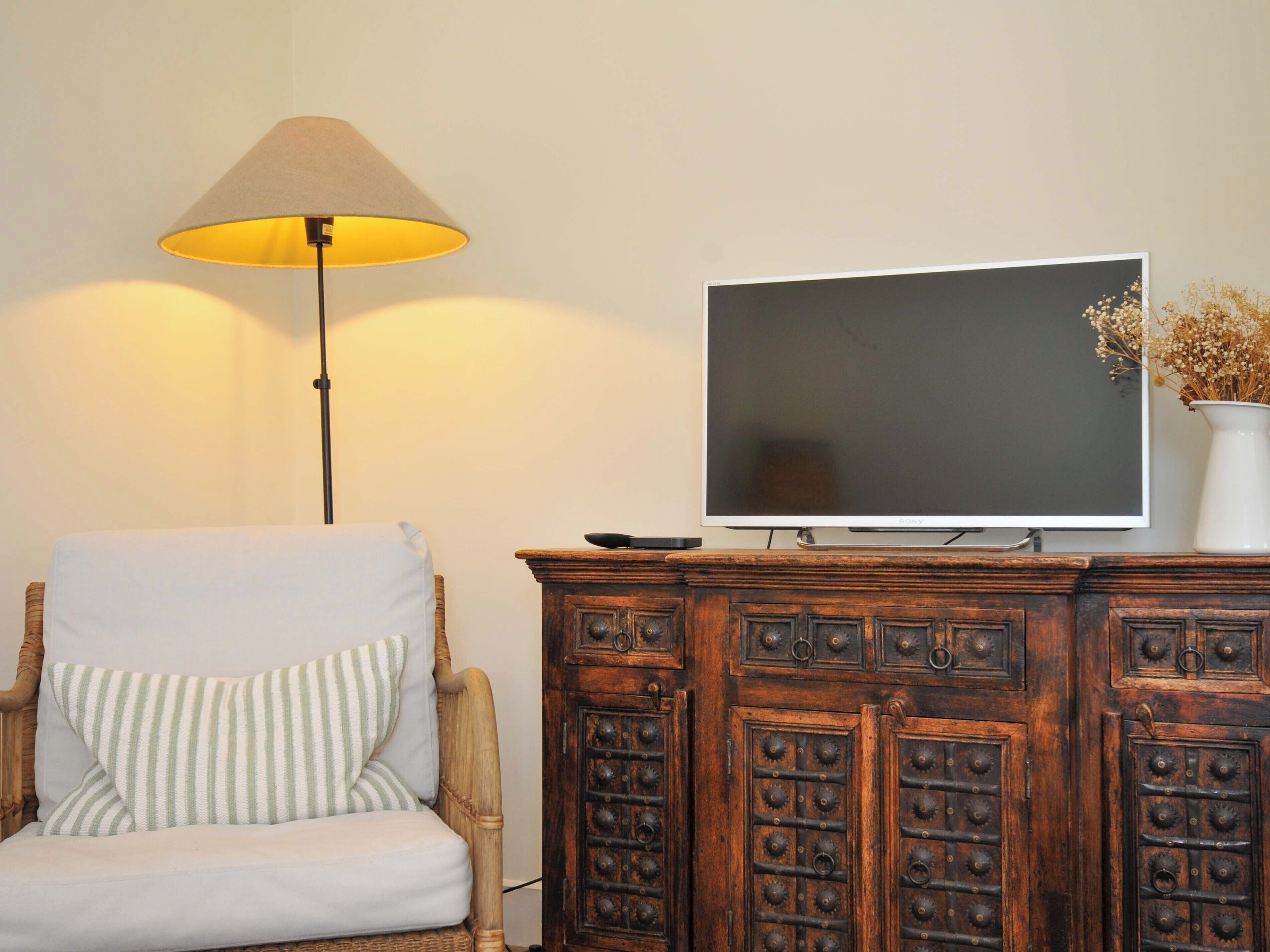 A room with a television on a wooden cabinet, a lamp, a wicker chair, and a vase with dried flowers.