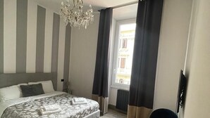 Makuuhuoneessa on sänky, pieni pyöreä pöytä, tuoli, kattokruunu ja ikkuna, jossa on verhot.