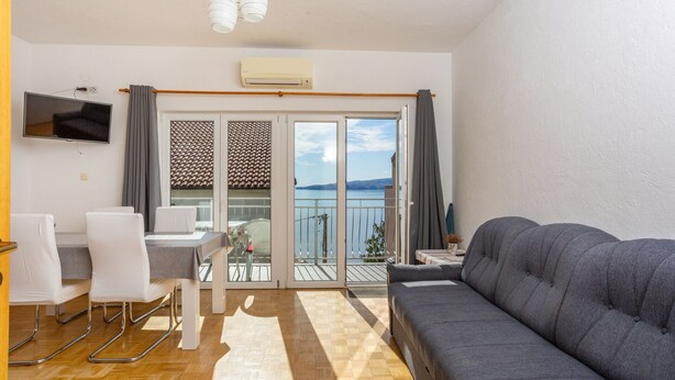 A living room with a sofa, a dining table with chairs, a TV mounted on the wall, and a balcony with a view of the water. - Charming Senj by Villas Guide (Senj)