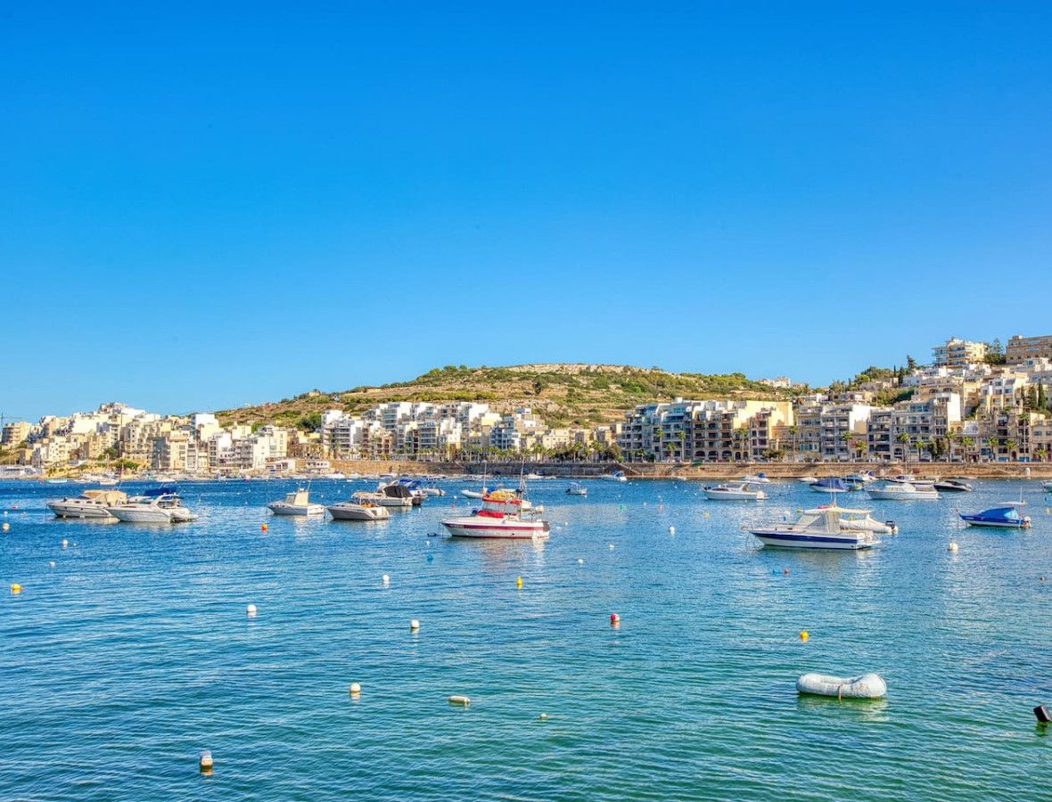 A coastal town with numerous boats docked along the waterfront, buildings on a hillside, and a clear blue sky.