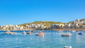 A coastal town with numerous boats docked along the waterfront, buildings on a hillside, and a clear blue sky.