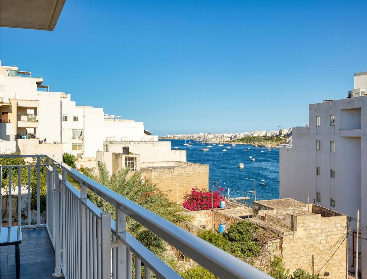 A coastal residential area with white buildings, a clear blue sky, and a view of the sea with boats.