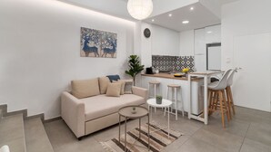 Een moderne woonkamer met een beige bank, een klein rond tafeltje, en een keukenhoek met een barkruk en een koelkast.