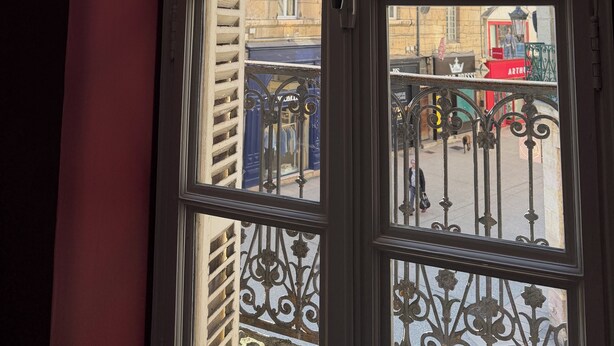 Blick durch ein Fenster mit einem Metallgitter und Gebäuden im Hintergrund.