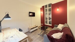 Ein modernes Zimmer mit einem Holztisch, einer schwarzen Schreibtischlampe, einem an der Wand montierten Fernseher, einem Glastisch, einer roten Wand und einem grauen Sofa mit weißer Decke.