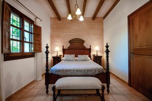 A bedroom with a wooden bed, two bedside tables with lamps, a bench, a large window, and a wooden door. - Exclusive Mallorcan finca with new house, pool and views of the countryside (PM)