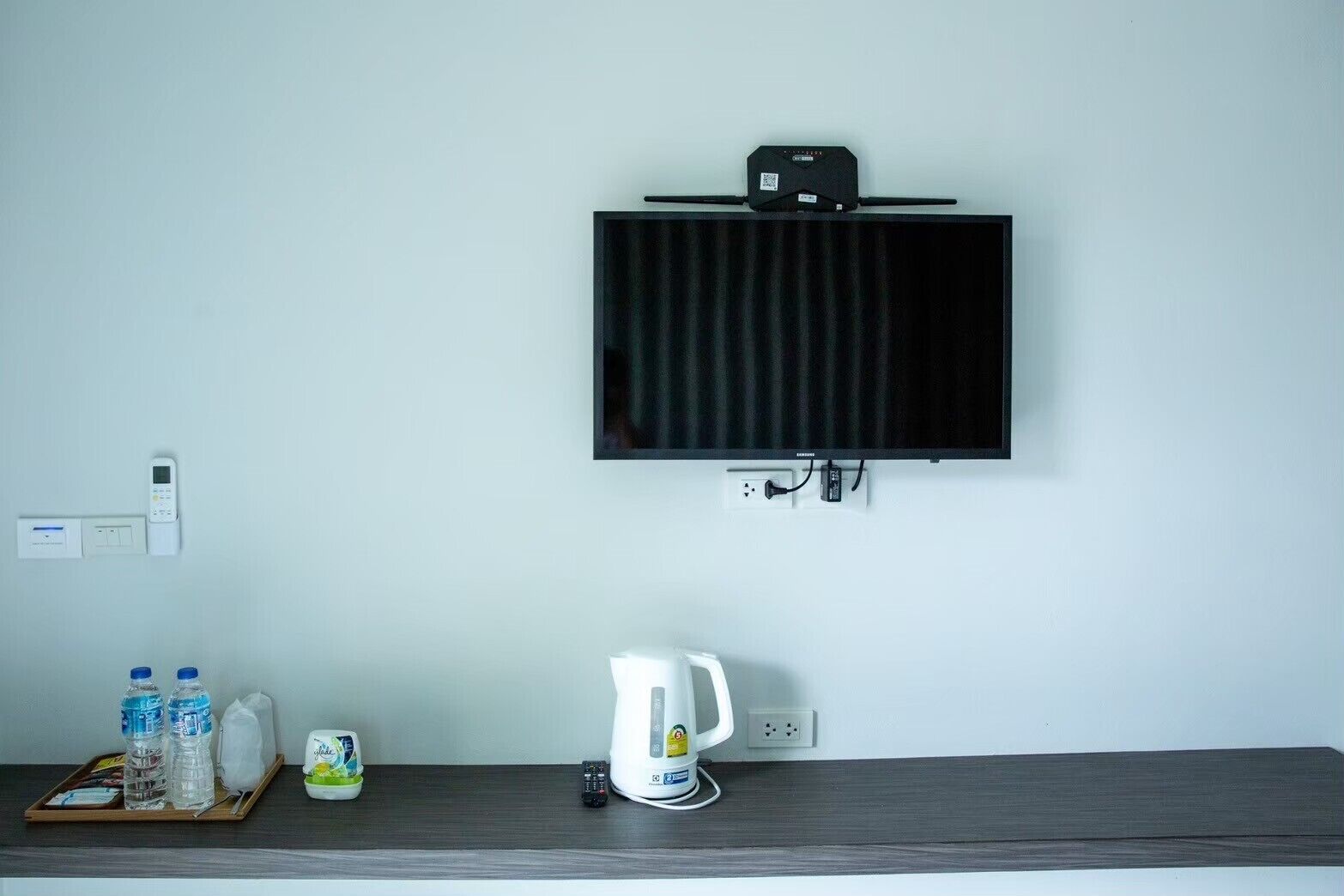 A hotel room with a mounted flat-screen TV, a thermostat, a kettle, and a tray with bottled water and snacks.