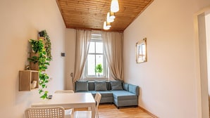 Een gezellige woonkamer met een houten plafond, een grijze hoekbank, een witte eettafel met stoelen en een potplant aan de muur.