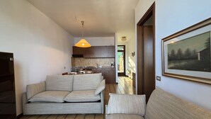Uma sala de estar moderna com sofá cinzento, piso de madeira e cozinha ao fundo.