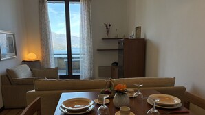 Uma sala de jantar com uma mesa posta para quatro pessoas, um sofá, uma luminária e vista para o exterior.