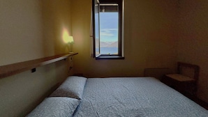 Um quarto pequeno e pouco iluminado, com uma cama de solteiro, uma mesa de cabeceira com abajur, e uma janela com vista para o exterior.