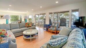 A spacious living room with a grey sofa, a round coffee table, and a blue patterned sofa. There is a television on the right and a large window providing a view of the outdoors.