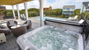 Un jacuzzi en una terraza de madera con mobiliario de mimbre, una piscina al fondo y casas a lo lejos.
