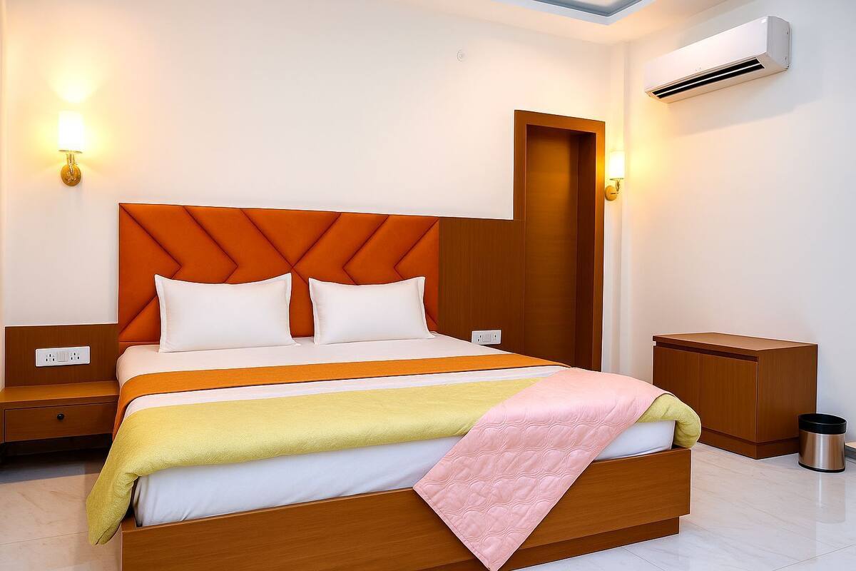 A modern hotel room with a large bed, a nightstand, a bedside table, an air conditioning unit, and a trash can.