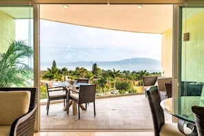 Een balkon met een tafel en stoelen, een glazen deur en uitzicht op zee en bergen.