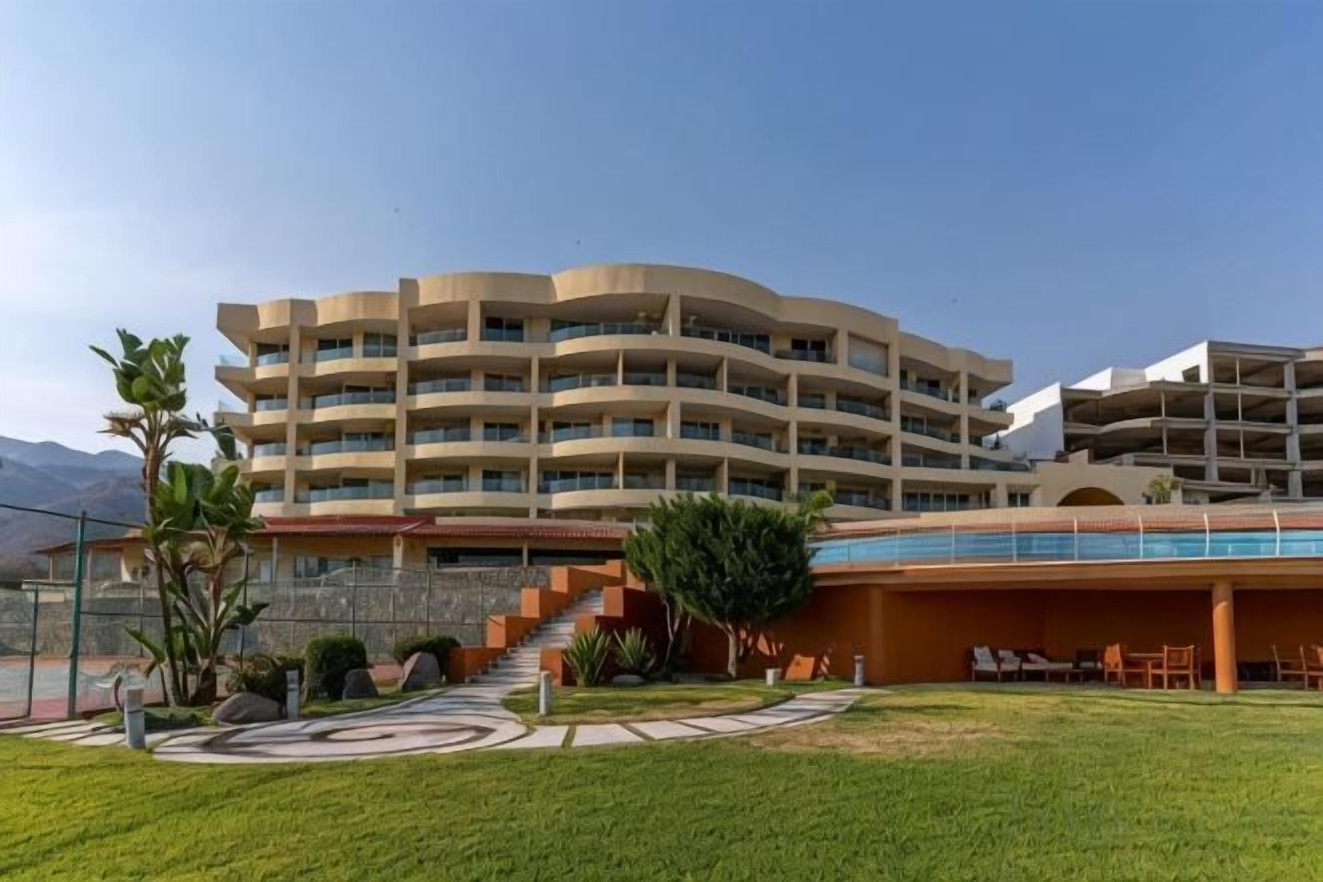 Un hotel de varios pisos con zona de piscina, mobiliario exterior y un jardín ajardinado.