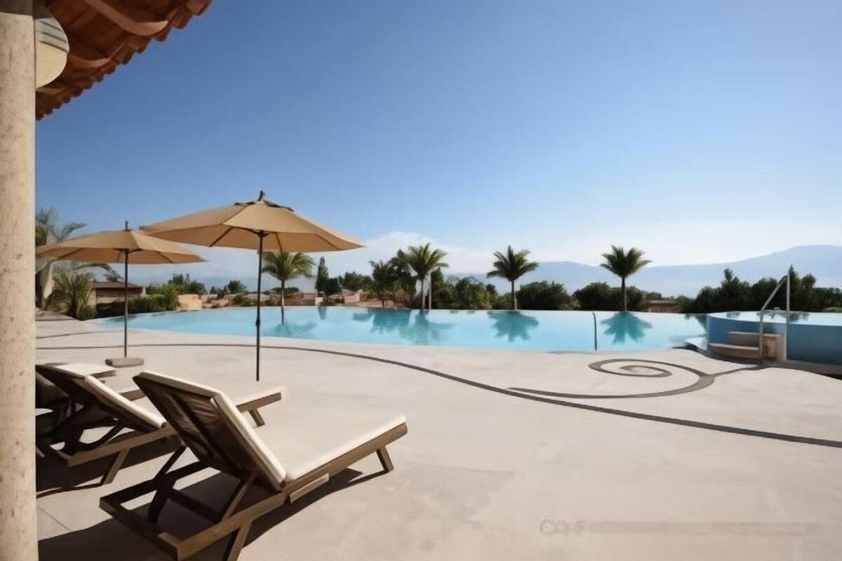 A pool area with lounge chairs and umbrellas, surrounded by palm trees and mountains.