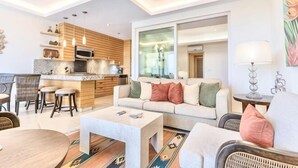 A modern living room with a sofa, coffee table, and dining area. There is a kitchen with wooden cabinets and a large window. - ANANA El Dorado 203 Condo with Lake views & Pool (Chapala)