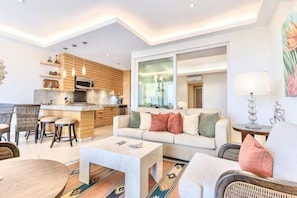Een moderne woonkamer met een bank, salontafel en een eethoek. Er is een keuken met houten kasten en een groot raam.