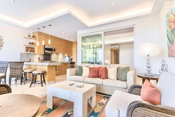 A modern living room with a sofa, coffee table, and dining area. There is a kitchen with wooden cabinets and a large window.