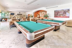 Une table de billard recouverte d’un drap de feutre vert, un canapé et un téléviseur dans une pièce dont le sol est carrelé et qui contient une bibliothèque.