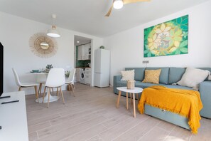 Una sala de estar moderna con un sofá, una mesa de comedor con sillas, una cocina al fondo y un cuadro abstracto grande en la pared.