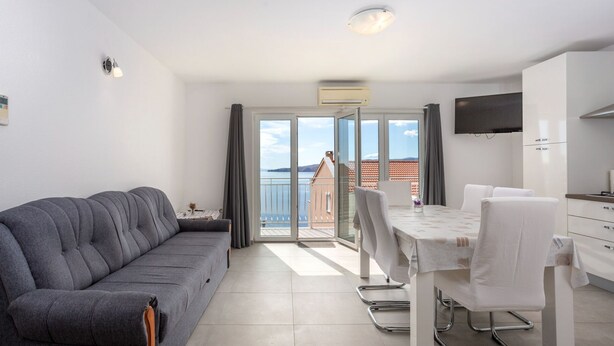 Ein modernes Apartment mit grauem Sofa, einem Esstisch mit Stühlen, einem an der Wand befestigten Fernseher und einem Balkon mit Meerblick.