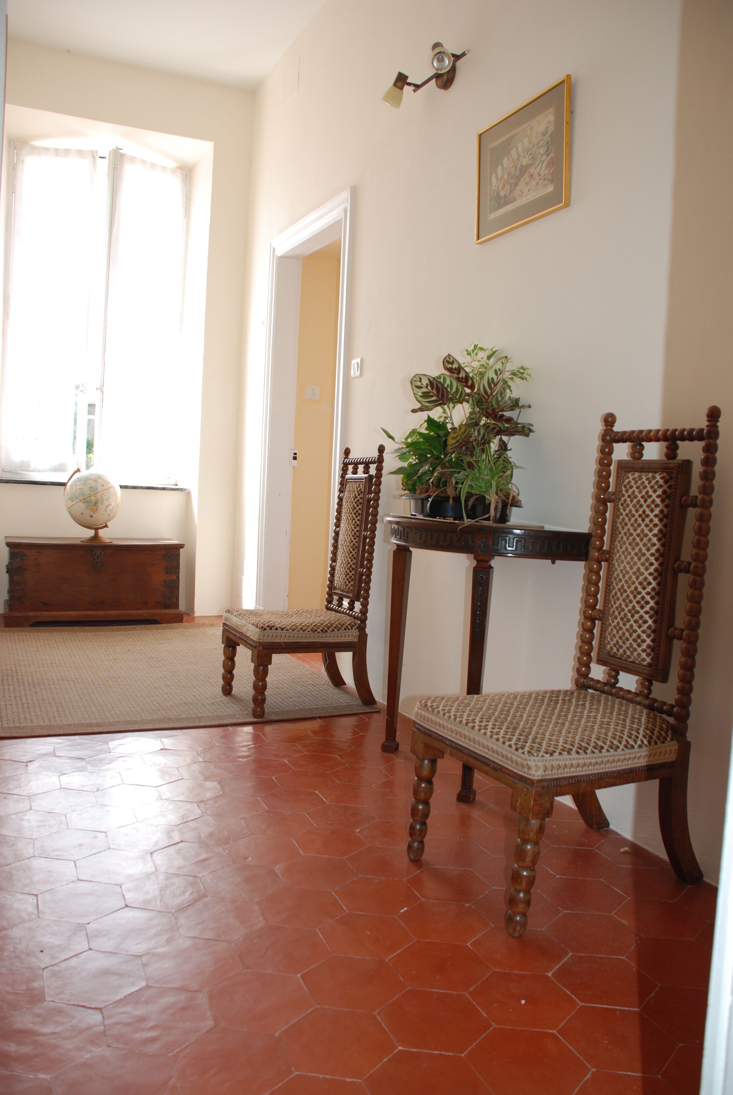 A room with a wooden floor, two wooden chairs, a globe on a wooden cabinet, a potted plant on a table, and a framed picture on the wall.