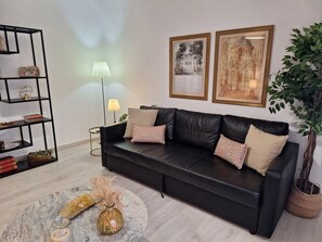 Ein schwarzes Ledersofa mit gemusterten Kissen, ein Marmortisch, eine Stehlampe, ein Bücherregal und zwei gerahmte Kunstwerke an der Wand.
