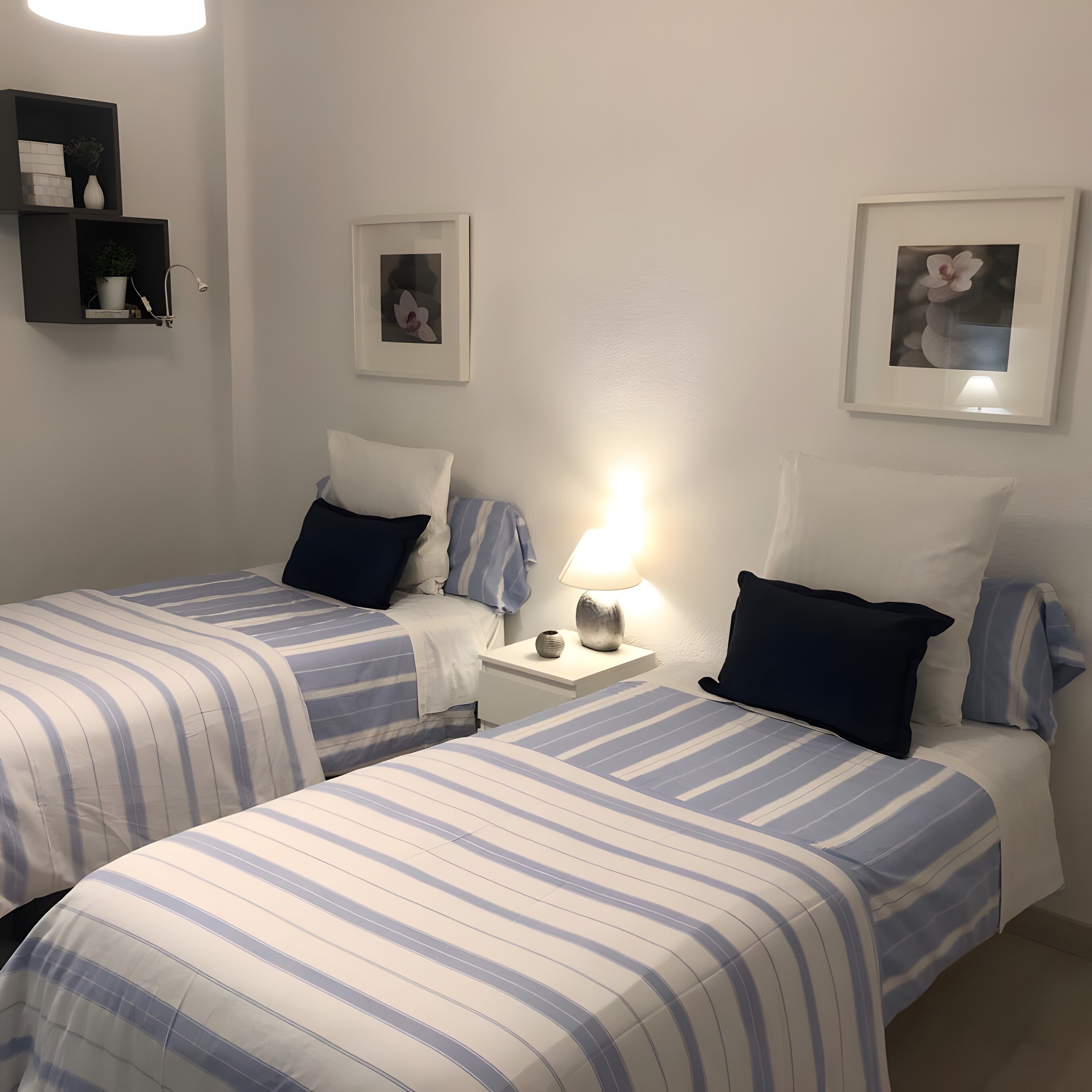 A hotel room with two beds, a bedside table with a lamp, a wall-mounted shelf with decorative items, and framed pictures on the wall.