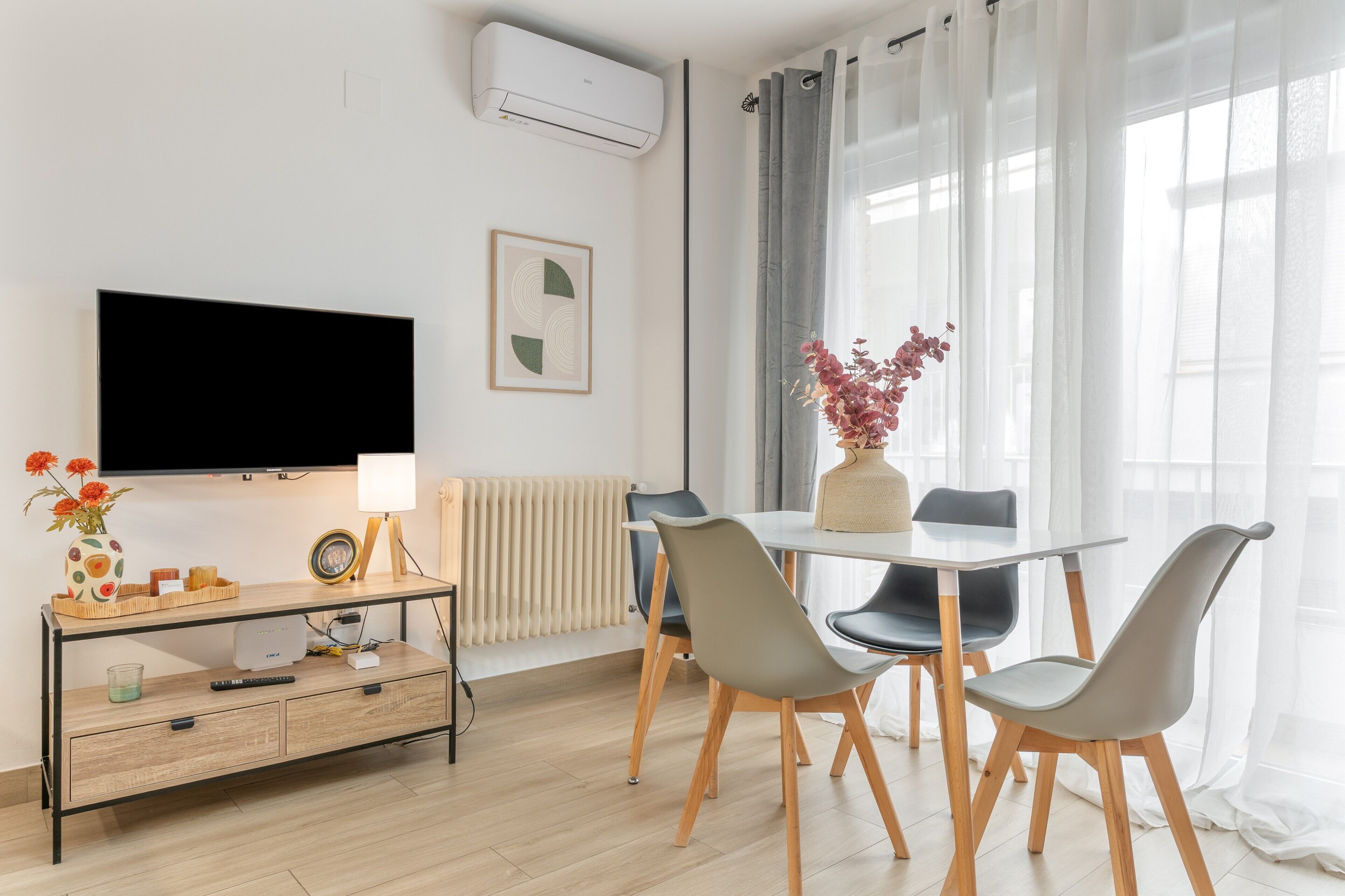 A modern living room with a flat-screen TV, a dining table with chairs, a small console table with a lamp, and a radiator.