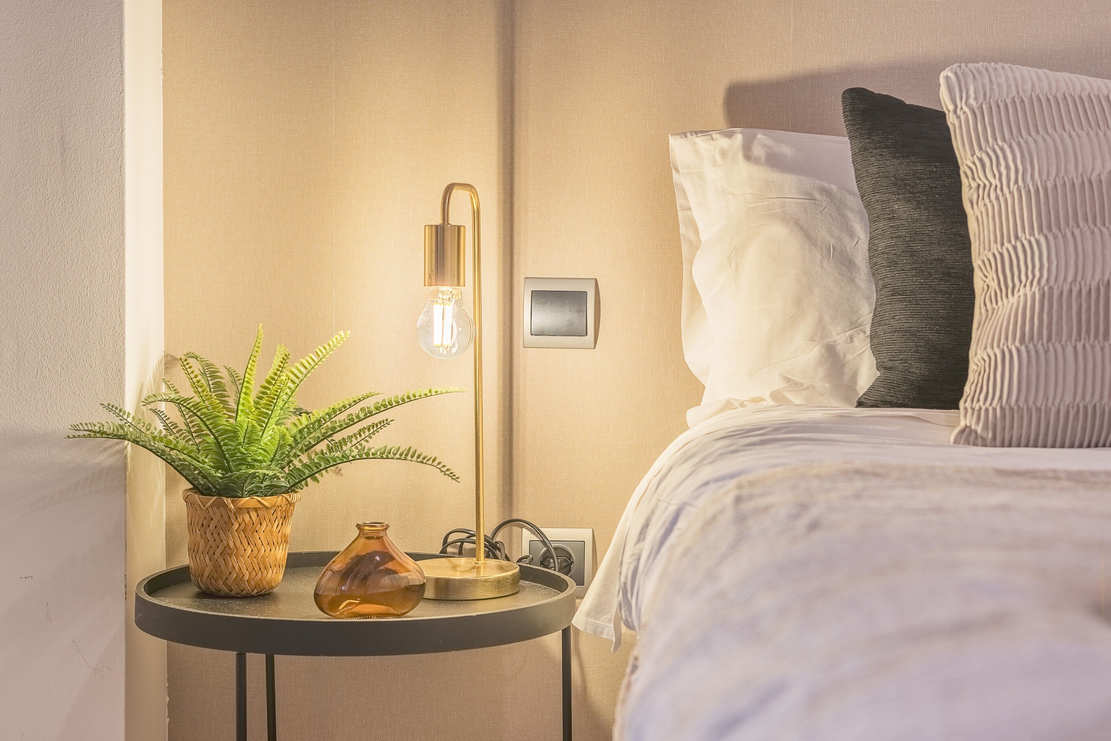 A neatly arranged bedroom with a bed, pillows, a bedside table with a lamp, a plant, and a vase.