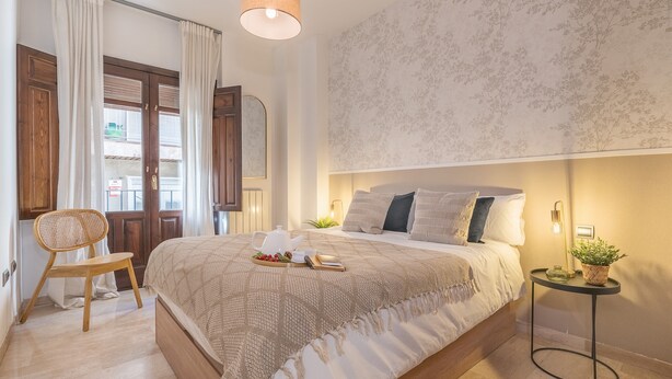 Ein Schlafzimmer mit einem Bett, einem Sessel, einem Nachttisch mit Pflanze und einer Wand mit floralem Muster.