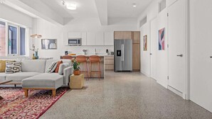 Un salon moderne avec un canapé, une table basse et un tapis à motifs. Il y a également un coin cuisine équipé d’un réfrigérateur, d’un four à micro-ondes et de tabourets de bar.