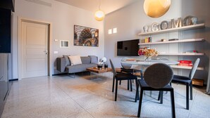 Ein modernes Wohnzimmer mit Sofa, Couchtisch und Essbereich. Ein Fernseher ist an der Wand montiert, darüber hängt ein Gemälde und Regale zeigen dekorative Gegenstände.