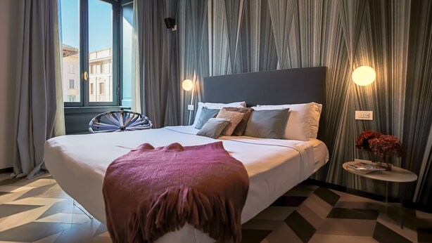 Ein Hotelzimmer mit einem großen Bett, einem runden Nachttisch mit einer Vase und Blick auf Gebäude durch das Fenster.